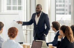 African american conference speaker coach talk to audience give presentation on flipchart to employees group, black trainer manager speaking training diverse corporate team at office meeting seminar